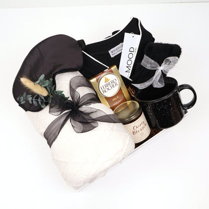Elegant sleep and loungewear gift box for her with black pajama set, quilted cream throw, ceramic mug, candle, and eye mask