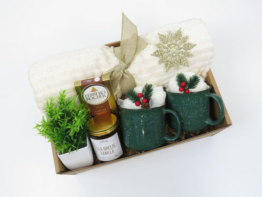 Festive holiday gift box with two patterned mugs, a scented candle, plush blanket, cozy socks, ornaments, and gourmet treats, packaged for family and friends.